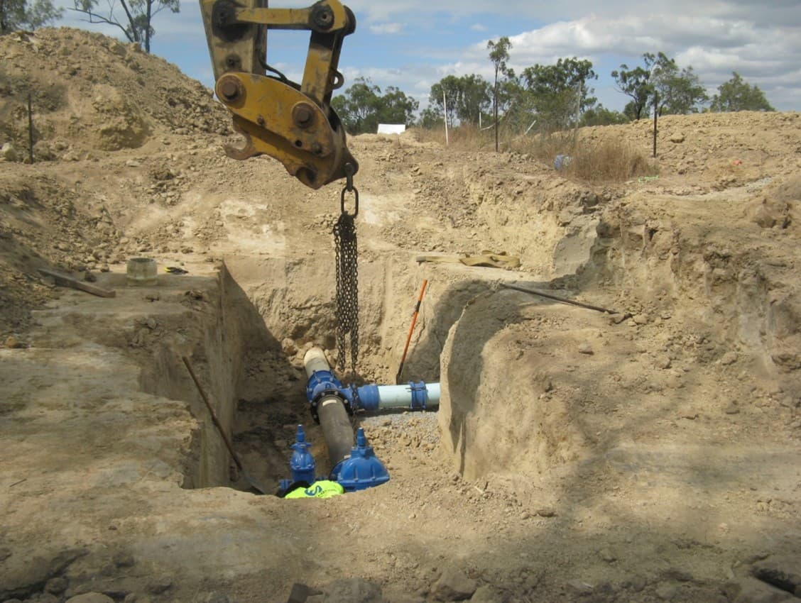 An excavator lowers pipe fittings into a deep, stepped trench on the Northshore sewerage works